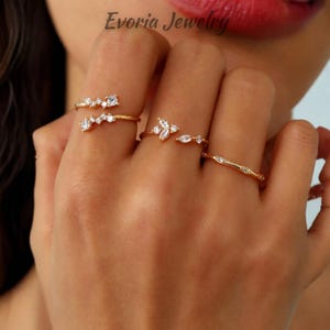 May include: Three gold-toned rings featuring clear, sparkling stones. The first ring has a cluster of small round stones, the second features a marquise and round stone arrangement, and the third is a thin band with embedded stones. Evoria Jewelry text.