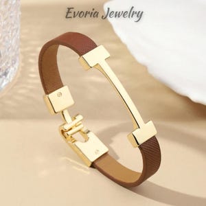 Peut inclure: Un bracelet en cuir texturé marron doté d'une pièce centrale en métal doré poli en forme de H et d'un fermoir en métal doré assorti. Le texte Evoria Jewelry est visible en haut de l'image, au-dessus du bracelet.