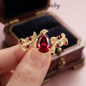 Può includere: Un anello in oro con una pietra preziosa rossa centrale a forma di pera, circondata da pietre rosse più piccole incastonate in una fascia decorata con motivi a foglia. La fascia in metallo ha una finitura lucida e presenta delicate foglie.