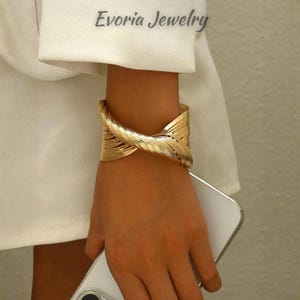 Puede incluir: Un brazalete tipo puño de tono dorado con un diseño trenzado de múltiples hilos y un acabado texturizado. El brazalete se lleva en la muñeca y el texto Evoria Jewelry es visible en la parte superior de la imagen sobre la manga blanca.