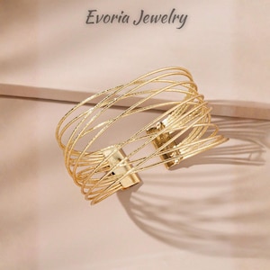 May include: A gold-toned cuff bracelet featuring multiple thin, textured wire strands that overlap in a crisscross pattern. The bracelet has a wide, open design with a polished metal clasp at the centre. The text Evoria Jewelry is visible.
