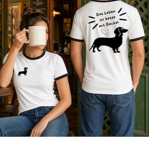 Könnte beinhalten: Ein weißes Ringer-T-Shirt mit schwarzem Besatz. Die Vorderseite zeigt eine kleine schwarze Dackel-Silhouette. Die Rückseite zeigt eine große schwarze Dackel-Silhouette, ein Herz-Symbol und den Text Das Leben ist beser mit Dackel.