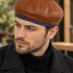 Puede incluir: Una boina de cuero marrón con un diseño estructurado de varios paneles y costuras visibles. La base de la gorra está acentuada con un borde de tela azul y tres pequeños remaches metálicos en el lateral, aportando un acabado detallado.