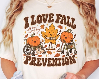 Könnte beinhalten: Ein elfenbeinfarbenes T-Shirt mit einer braunen Grafik und dem Text I LOVE FALL PREVENTION. Das Design zeigt einen lächelnden Kürbis im Rollstuhl, ein lächelndes Ahornblatt und einen lächelnden Kürbis mit Rollator, umgeben von herbstlichen Blättern.