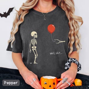 Peut inclure: Un t-shirt gris anthracite à manches courtes avec un motif représentant un squelette face à une main squelettique tenant un ballon rouge. Le texte "Well, shit...." est imprimé en blanc sous le ballon. Le t-shirt a un col rond.