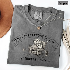 May include: A pepper-coloured t-shirt featuring a graphic of a toad sitting in an armchair with a cup, accompanied by the text "WHAT IF EVERYONE ELSE IS? JUST UNDERTHINKING?" The shirt has a crew neck and a Comfort Colors brand label at the collar.