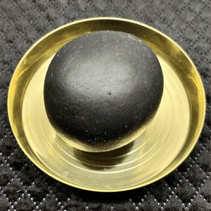 Pode incluir: Um objeto de pedra preto fosco, liso e arredondado, centralizado dentro de uma bandeja circular de metal dourado polido. A bandeja apresenta uma série de ranhuras circulares concêntricas gravadas na base, criando um contraste com a pedra.