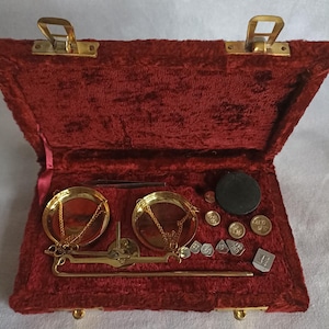 Könnte beinhalten: Eine goldfarbene Balkenwaage mit zwei runden Waagschalen und einem zentralen Balken, untergebracht in einem rechteckigen Etui mit tiefrotem Samtfutter. Das Set enthält verschiedene kleine Metallgewichte, eine schwarze Scheibe und eine Pinzette.