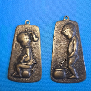 May include: Two rectangular bronze-toned metal plaques with rounded corners and top loops. The left plaque features a relief of a child sitting on a chamber pot. The right plaque features a relief of a child standing and using a chamber pot.