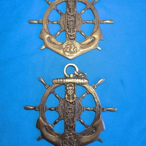 Könnte beinhalten: Zwei identische Wandplaketten aus Messing, die ein Schiffssteuerrad kombiniert mit einem vertikalen Ankerdesign zeigen. Jedes Stück verfügt über einen zentralen Anker, ein kleines Steuerrad-Emblem und ein Relief eines Segelschiffs am Ankerfuß.