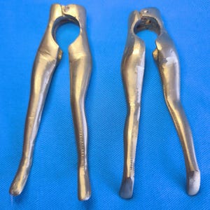 Puede incluir: Dos cascanueces de metal de color dorado, cada uno con un diseño en forma de par de piernas. Los cascanueces tienen un mecanismo de bisagra en la parte superior, lo que permite que los dos mangos en forma de pierna se abran y cierren.