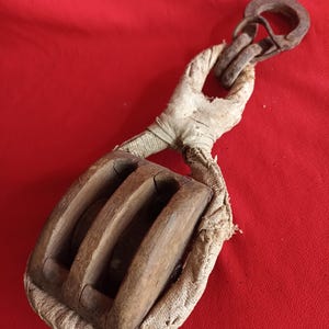 Könnte beinhalten: Ein alter Holzflaschenzug mit Doppelscheibe, eingefasst in verwittertes Holz. Der Flaschenzug ist mit einem dicken, ausgefransten, cremefarbenen Canvas-Band befestigt, das durch einen rostigen Eisenhaken an der Oberseite geführt wird.