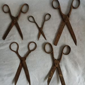 May include: A collection of five vintage metal scissors with a weathered, rusted brown patina. Each pair features two circular finger loops and two blades connected by a central pivot point, displaying varying sizes and blade shapes throughout.