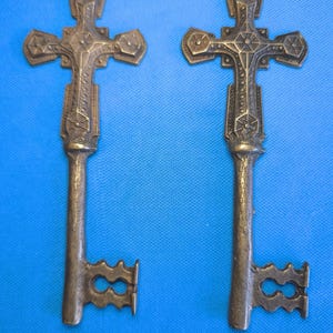 May include: Two identical bronze-colored metal keys featuring an ornate cross design at the bow. The cross has a textured, geometric pattern with small circular accents. The shank is cylindrical with a rough, hammered texture, and the bit is shaped.