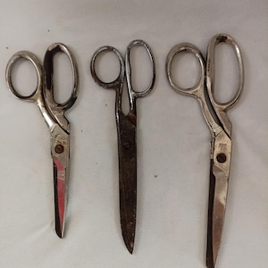 May include: Three pairs of metal scissors arranged side by side. The left pair is short with a shiny finish, the middle pair is long with a dark, weathered patina, and the right pair is long with a polished silver-tone finish and a central screw.