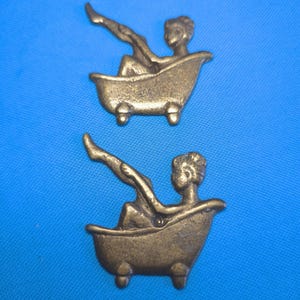 Puede incluir: Dos adornos de metal idénticos en tono latón, cada uno representa una figura sentada en una bañera con patas con una pierna levantada. Las figuras tienen el pelo corto y están orientadas hacia la derecha sobre cuatro pies.