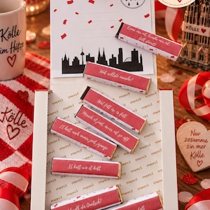 May include: A white gift box containing 16 chocolate bars with red wrappers featuring German text and confetti patterns. The box lid displays a black city skyline silhouette, a coat of arms, and the text 16 Stück. The interior base is printed with the word merci.