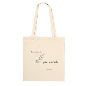 Könnte beinhalten: Eine cremefarbene Stofftasche aus Canvas mit zwei langen Tragegriffen. Auf der Vorderseite befindet sich eine kleine grüne Strichzeichnung eines Zweigs sowie der deutsche Schriftzug "Ich mach das jetzt einfach" in dunkelgrüner Serifenschrift.