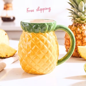 Peut inclure: Une tasse en céramique en forme d'ananas avec un corps jaune texturé, un bord et une anse verts. La tasse présente une surface à motif en losanges imitant la peau d'un ananas, avec une finition lisse et brillante sur toute la pièce.