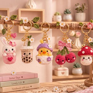 May include: Five crochet keychains hanging from a wooden branch, featuring a white bunny in a pink cup, a boba tea, a yellow chick in a purple hat, two red cherries with a pink bow, and a red mushroom with white spots, all with gold rings.