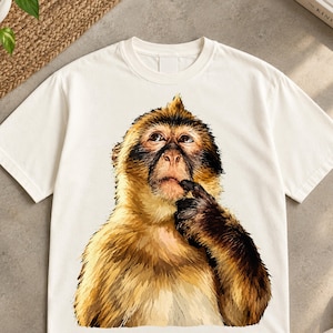 May include: A cream-colored short-sleeve t-shirt featuring a large, detailed graphic print of a brown monkey with a tuft of hair on its head. The monkey is depicted with its hand raised to its chin in a thoughtful pose, looking upward with brown eyes.