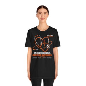Könnte beinhalten: Ein schwarzes T-Shirt mit einem orangefarbenen Stethoskop in Herzform und einer EKG-Linie in der Mitte. Der Text lautet JUVENTA HEALTHCARE SERIES, MENSCHEN HELFEN. MACHT DEN UNTERSCHIED. sowie EMPATHIE, PFLEGE, STÄRKE, TEAMGEIST in Orange und Weiß.