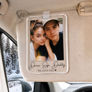 Puede incluir: Un clip rectangular de acrílico transparente para el parasol del coche con una fotografía impresa de una persona con cabello largo castaño y otra persona con gorra de béisbol. El texto "Drive Safe, Daddy" y "WE LOVE YOU" con un pequeño icono de corazón están impresos debajo.