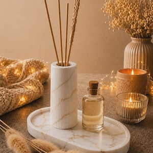 Peut inclure: Un plateau ovale en marbre blanc avec des veines dorées supporte un diffuseur cylindrique cannelé avec quatre bâtonnets et une petite bouteille en verre transparent avec un bouchon en liège, accompagné d'herbes séchées et d'un éclairage chaleureux.