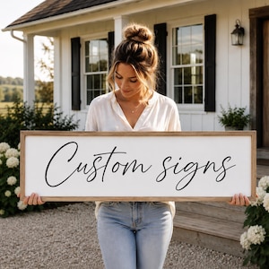 May include: A rectangular wooden sign with a white background and a natural wood frame. The text 'Custom signs' is written in a black, flowing cursive script across the center of the sign. The sign is held horizontally by a person.