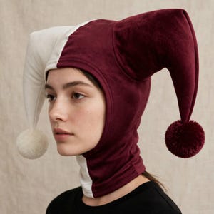 Pode incluir: Um chapéu de bobo da corte de dois tons feito de tecido de veludo, com um design dividido em creme e bordô profundo. O chapéu tem duas longas extensões laterais pontiagudas, cada uma terminada com um pompom correspondente na ponta.