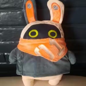 May include: A plush toy shaped like a rabbit with long ears, featuring a cream-colored body, a dark gray jacket, and a bright orange bandana. The rabbit has a black face mask with two circular yellow eyes and silver accents on the ears.