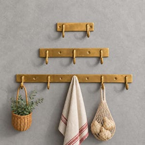 May include: Three horizontal brass wall racks with varying numbers of hooks. The top rack has two hooks, the middle has three, and the bottom has six. A small woven basket with greenery and a beige towel with red stripes hang from the bottom rack.