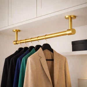 Puede incluir: Una barra de armario de metal dorado montada en la parte inferior de un estante blanco. La barra cuenta con dos soportes cilíndricos de techo y sostiene cinco perchas de plástico negro, cada una con un blazer en colores beige, verde, azul y negro.