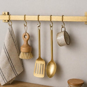 May include: A brushed gold metal wall-mounted rail featuring five matching S-hooks. Hanging from the hooks are a beige striped linen towel, a wooden dish brush, a gold metal slotted spatula, a gold metal spoon, and a small speckled ceramic cup.