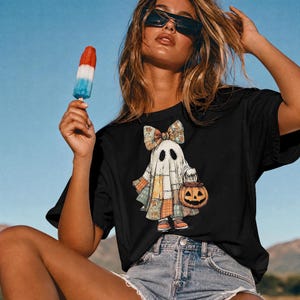 Könnte beinhalten: Ein schwarzes T-Shirt mit einer Grafik eines Geistes, der eine gemusterte Schleife trägt und einen Kürbis-Eimer hält. Der Geist ist in einem Patchwork-Stil in gedeckten Orange-, Blau- und Cremetönen dargestellt und trägt orangefarbene Schuhe.