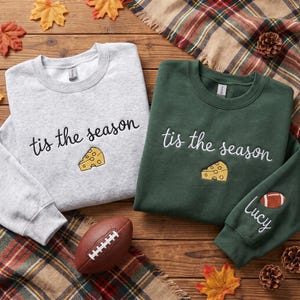May include: A light gray crewneck sweatshirt and a dark green crewneck sweatshirt, both featuring the embroidered text 'tis the season' in cursive and a yellow Swiss cheese wedge. The green sweatshirt also has a football and the name 'Lucy'.
