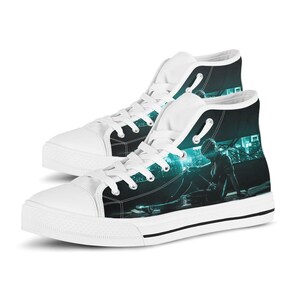 Può includere: Un paio di scarpe alte con punta in gomma bianca, lacci bianchi e tomaia in tela nera stampata con una grafica color ottanio e nero raffigurante una figura in un ambiente futuristico. Le scarpe hanno una suola bianca con una riga nera.