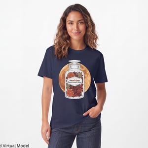 Può includere: Una t-shirt blu navy con la grafica di un barattolo da speziale in vetro riempito con fette d'arancia essiccate, bastoncini di cannella e anice stellato. L'etichetta riporta la scritta Warm & Cosy Home Sweet Home su una luna dorata.