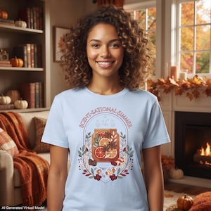 Può includere: Una t-shirt azzurra con la grafica di un barattolo di vetro riempito con fette d'arancia, bastoncini di cannella e anice stellato. Il testo SCENT-SATIONAL SIMMER è stampato ad arco sopra il barattolo, circondato da illustrazioni botaniche.