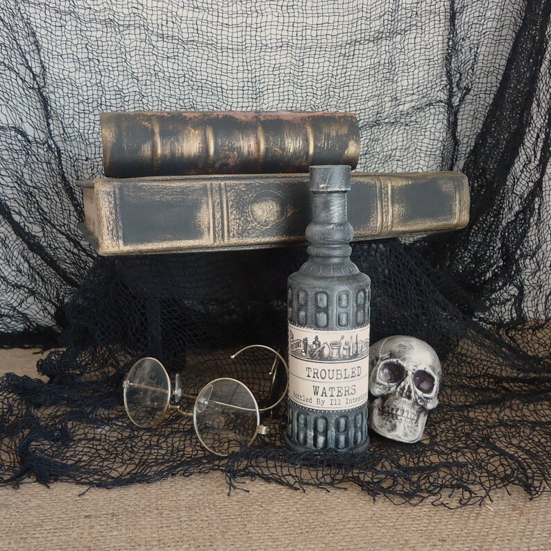 May include: A still life arrangement featuring a gray bottle labeled "Troubled Waters" with a skull, antique books, and round eyeglasses. The bottle is flanked by a skull and vintage books, all set against a black net.
