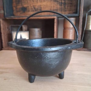 May include: A black cast iron cauldron with a textured surface, featuring three short legs, a wide circular opening, and a thin, arched metal handle attached to two side lugs. The vessel has a deep, rounded bowl shape and a sturdy, heavy appearance.