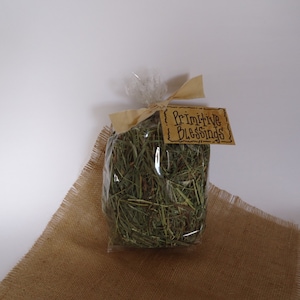 May include: A clear plastic bag filled with dried green grass, tied with brown paper and a brown tag that reads "Primitive Blessings". The bag is sitting on a brown burlap surface.