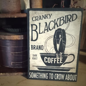 May include: A rectangular sign with the text "Cranky Blackbird Brand Dark Roast Coffee" and an illustration of a black bird inside a coffee cup. The sign also reads "Something to Crow About."