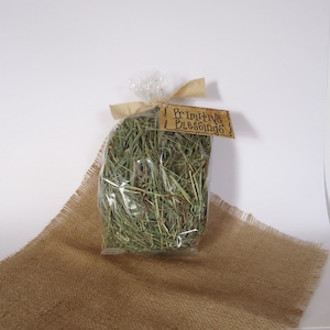 May include: A clear plastic bag filled with dried green grass, tied with a brown ribbon and a brown paper tag that reads "Primitive Blessings". The bag is sitting on a brown burlap surface.