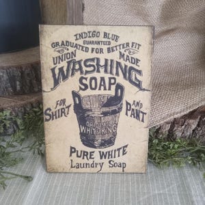 May include: Vintage-style sign advertising "Washing Soap" for shirts and pants. The sign features a black and white illustration of a bucket with the text "Original White King" and "Pure White Laundry Soap".
