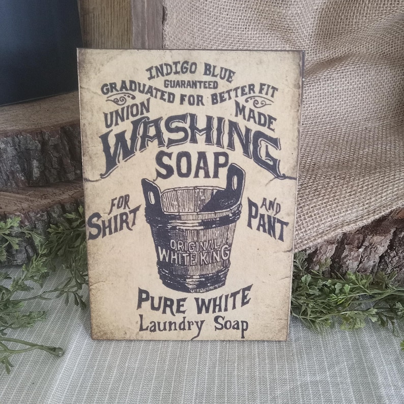 May include: Vintage-style sign advertising "Washing Soap" for shirts and pants. The sign features a black and white illustration of a bucket with the text "Original White King" and "Pure White Laundry Soap".