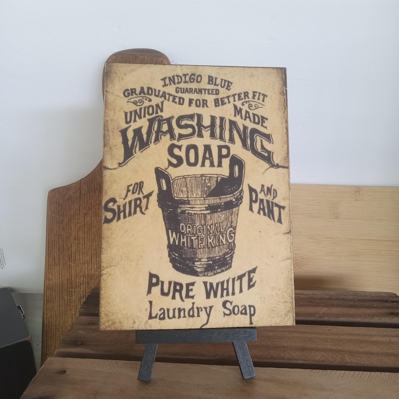 May include: A vintage-style sign advertising "Indigo Blue" washing soap. The sign features a bucket with the text "Original White King" and the words "For Shirt and Pant Pure White Laundry Soap".