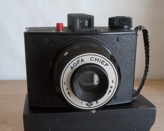 May include: A black Agfa Chief box camera featuring a textured body, a central lens with a white circular faceplate, a red shutter release button on top, a black viewfinder, and a black braided wrist strap attached to the side of the camera housing.