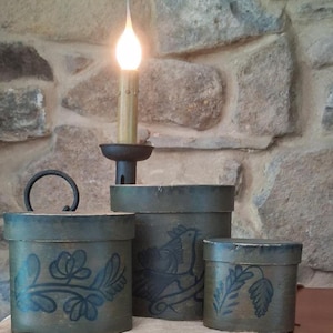 May include: Three cylindrical olive green wooden boxes with lids, each featuring dark blue painted botanical and bird motifs. A black metal candle holder with a lit flame stands behind the central box, which has a small black metal handle.