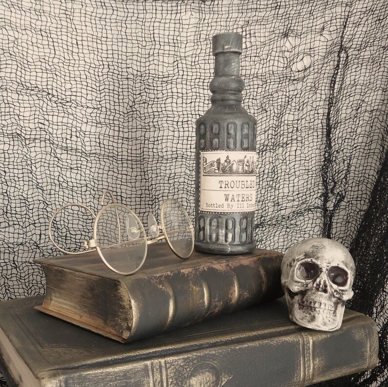 May include: A vintage-style scene featuring a gray bottle labeled "Trouble Waters" with a decorative label, a pair of round eyeglasses, a skull, and two antique books. The background is a black and white mesh.